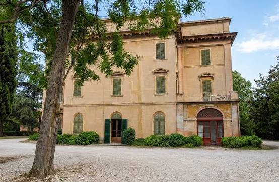 Facciata di Villa Fontedamo: palazzo ottocentesco color ocra con persiane verdi, dietro un grande albero.